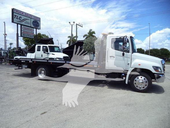 Tow truck with long wheelbase, white cab, flatbed, TLC Truck Sales Center sign in background. Visible text on sign includes TLC and EVTV logos. No other legible labels or serials.