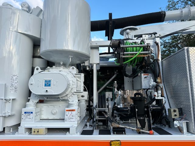 Industrial machinery compartment showing large white pump, control panel, hydraulic lines, and green hose in a truck equipment bay.