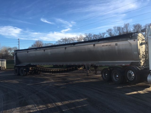 Trailer side view showing a second trailer with cylindrical aluminum tank, wheels at rear, blue sky and trees in background.