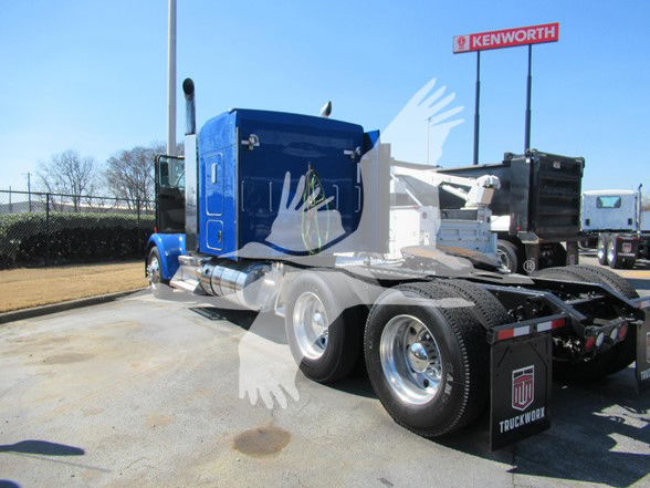 Kenworth W900L truck tractor with blue cab and dual rear axles, seen in a dealership lot. Visible text includes Kenworth sign in background and trailer undercarriage details.