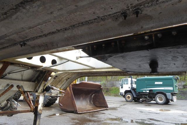 Underbelly of a Fruehauf dry bulk trailer showing underside beams, landing gear, and a nearby construction bucket and truck in background.
