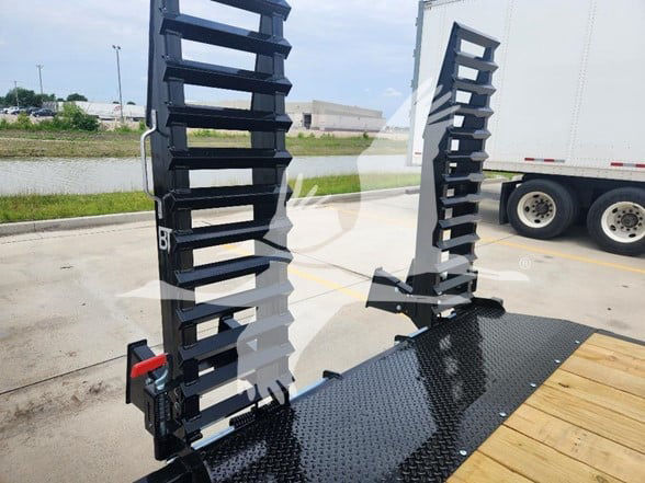Flatbed trailer accessory posts with ladder-like upright stakes on a black diamond-plate deck, with a white trailer in the background.