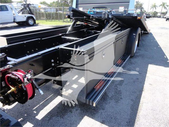 Tow truck chassis with hydraulic underlift components and toolboxes, black paint, red hose reel at front left, side steps and storage compartments visible.