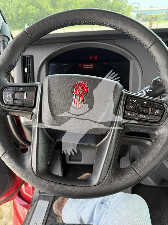 Steering wheel of a Kenworth truck interior with KW emblem and controls on both sides, digital gauge cluster in background.