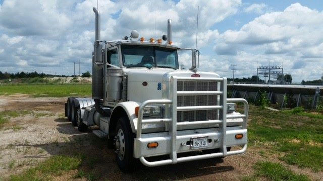 Peterbilt 379 conventional daycab truck in a field, white exterior, grille guard, stacked exhaust stacks, 3-axle tractor with front bumper and chrome accents.