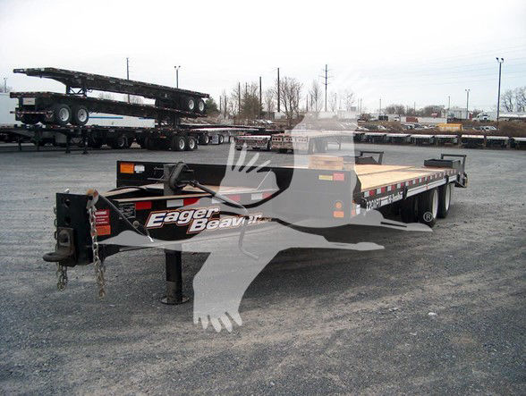 Flatbed trailer in a dealership lot showing Eager Beaver logo on front with black frame and wooden deck, gooseneck hitch, and spare tires in background.