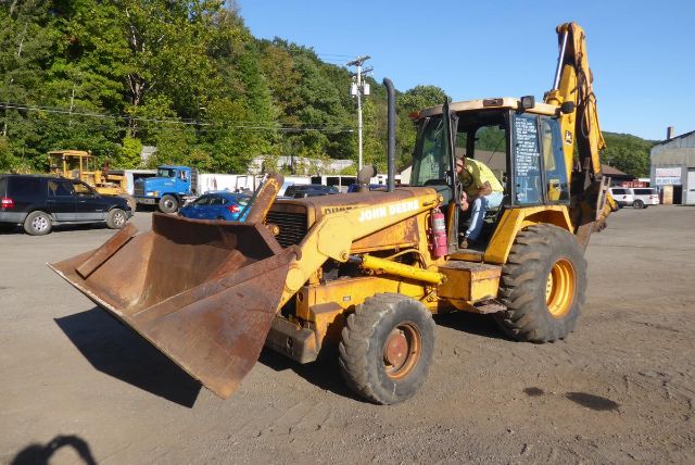 Backhoe loader with front bucket and rear digging arm, yellow John Deere 710B in a used equipment lot.