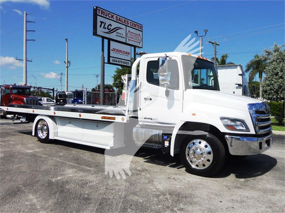 Tow truck with white cab and flatbed, sign in background reading TLC Truck Sales Center, chrome wheels and mirrors visible, blue sky.