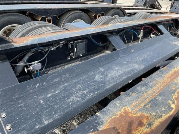 Trailer component area showing axles and undercarriage with hoses, wiring, and metal frame surface. Visible rust on edge plates and a black metal deck.