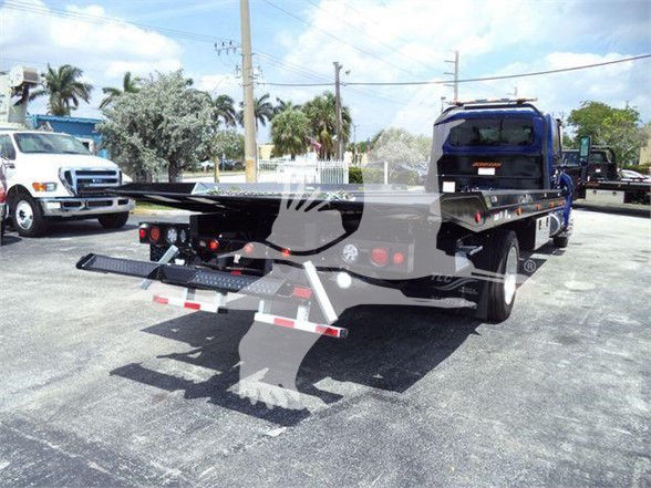 Truck bed with hydraulic rollback tow in a parking lot showing ramps and green vegetation in background.