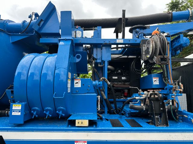 Blue Mack vacuum truck equipment with reels, hoses, and hydraulic components. Visible labels and warning stickers on metal frame.