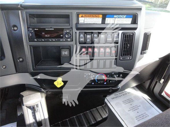 Cockpit of a Mack MD6 truck showing instrument cluster, radio, climate controls, and various switches. Visible labels include WARNING and NOTICE stickers, and a yellow hood release handle.