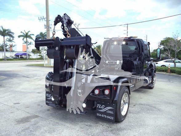 Tow truck from rear-right side showing hydraulic crane/tower with grapple and hoist, International MV cab in background, painted dark color with reflective decals.