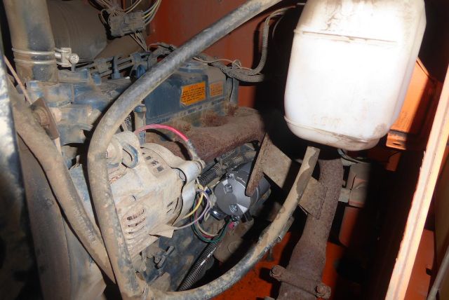 Trucking equipment engine bay showing alternator, wiring, hoses, and a white reservoir; compact diesel engine with orange machinery in background.