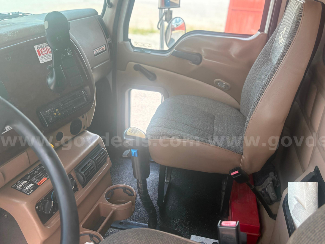 Cab interior of a 2002 Mack CV713 truck showing tan and brown dashboard, steering wheel, center console, manual gear lever, and driver seat with fabric/vinyl trim.