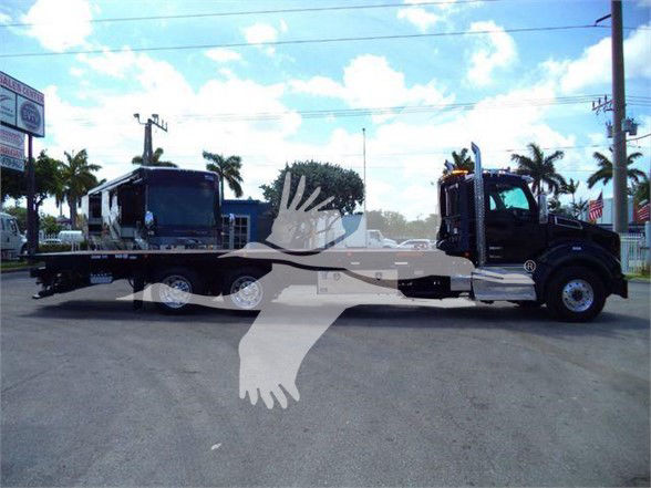 Kenworth T880 tow truck tractor with a flatbed trailer, black cab, on a daylight lot with palm trees in background.