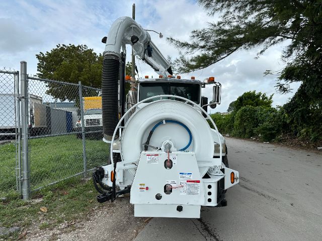 Vehicle back view of a white Peterbilt vacuum truck with hose reel and rear equipment, chain-link fence, trees, and a cloudy sky.