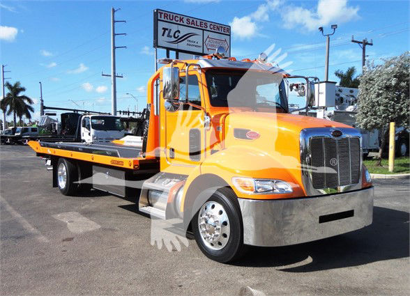 Tow truck with bright orange Peterbilt 337 cab and flatbed, chrome bumper, in a dealership lot.