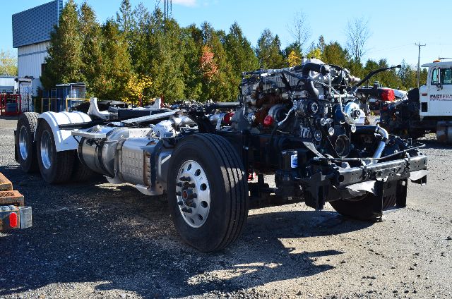 Front-left view of a cab-over truck chassis with engine and transmission exposed, showing a large exposed engine bay and dual rear axles.