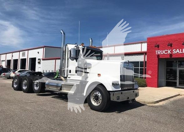 Kenworth W900L conventional daycab truck in white, parked outside a dealership, with chrome exhaust stacks and dual rear axles.