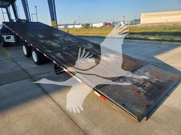 Flatbed trailer with a lowered ramp, used condition, showing metal surface and wheels along the side.