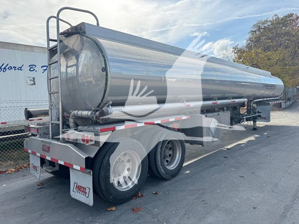 Tank trailer with a polished stainless steel cylindrical tank and rear wheels visible; ladder at rear, pump off-line configuration, Heil branding on mudflaps.