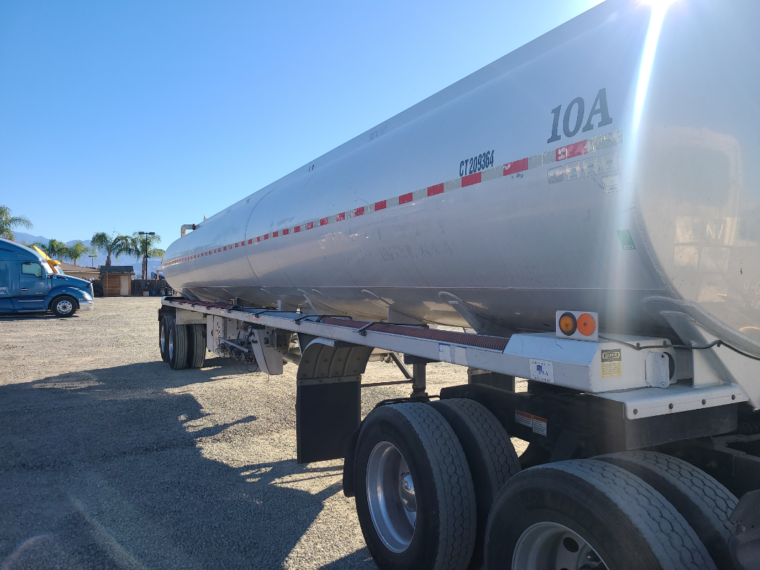 Fuel tanker trailer side view showing tandem axles and reflective red/white striping, 10A marking near front, with blue sky and other truck in background.