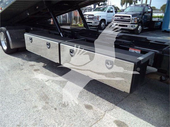 Three silver rectangular under-bed toolboxes mounted on a trailer frame under a raised bed, with handles on each toolbox; in background several trucks parked.