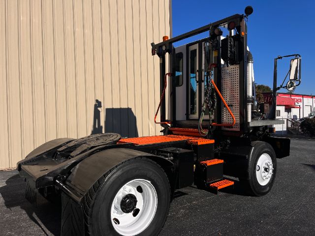 Industrial yard truck chassis with orange steps and a black metal frame, cab area with door, and single rear axle. Flatbed rear, white wheels, blue sky.