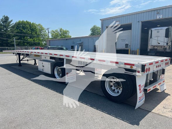 Flatbed trailer in a truck yard showing tandem rear axles, white wheels, and reflective tape along the sides with rear fender skirts.