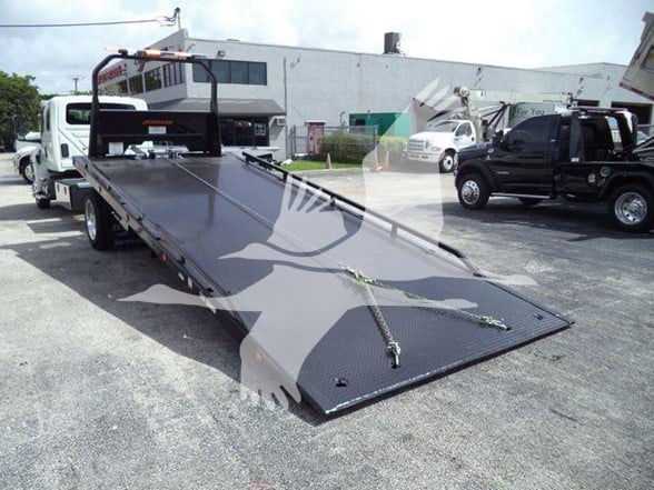Flatbed tow truck with a long black steel ramp trailer on a dealership lot, side rails, chains, and hook points visible.