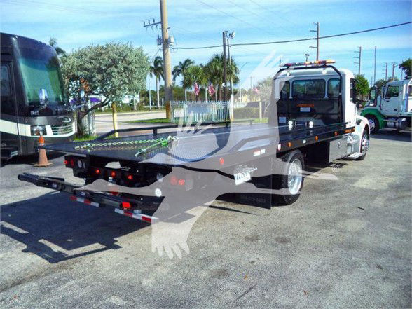 Tow truck on a lot, Peterbilt 536 flatbed with a long wheelbase and rear hitch, side mirrors, orange clearance lights, and light bar on cab roof.