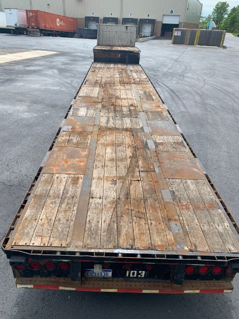 Flatbed trailer long wooden deck with weathered planks, rear lights, and metal framing. Orientation is sideways in the image with a cab and buildings in the background.