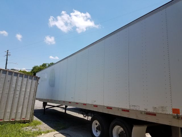 Trailer side view showing a large white utility freight trailer with riveted metal panels and support wheels; blue sky with a few clouds.