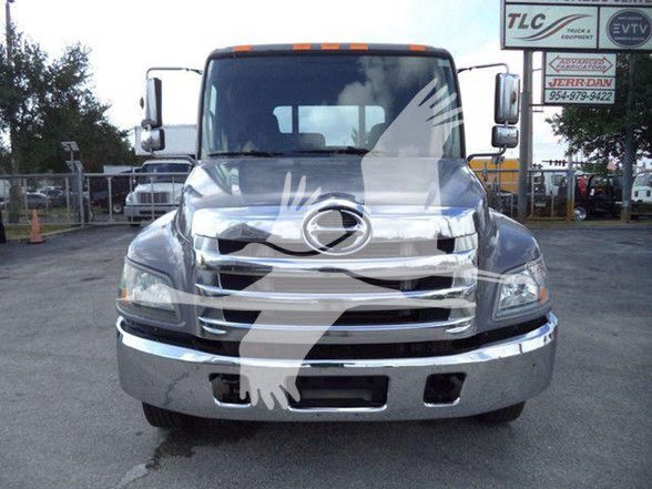 Front view of a 2014 Hino 258ALP tow truck with chrome bumper and a large Hino emblem on the grille. The truck has a damaged front end with visible chrome grille bars and a blank license plate area.