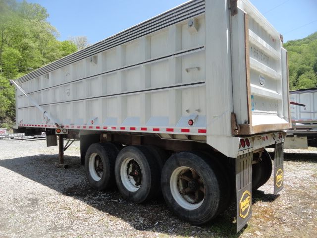 Trailer side view showing white dump trailer with triple axle and rear gate, with wheels and mud flaps visible.