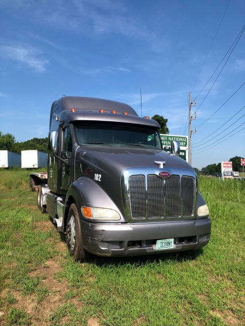 Peterbilt 387 truck front view, dark gray/blue cab, chrome grille, orange marker lights, Florida dealer signs in background, license plate visible, wheel and tire on left, grassy lot.