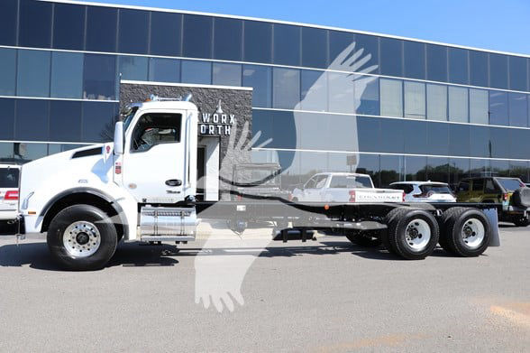 Kenworth T880 cab and chassis truck in a dealership lot, white cab, long wheelbase chassis with dual rear axles, reflective building in background.