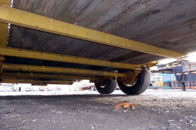 Underbelly of a trailer showing dual wheels, yellow frame rails, and wooden deck. Ground with small pebbles; a small orange object near the wheels.