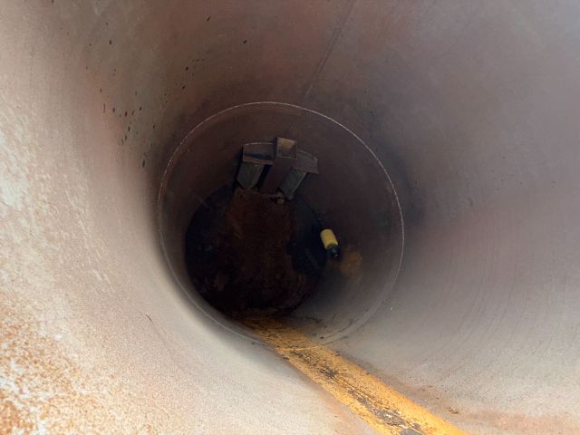 Internal view of a vertical cylindrical duct or chute with a dark interior, showing a narrow yellow-brown residue line along the bottom and some metal brackets inside.
