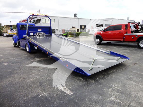 Tow truck with a blue flatbed ramp shown in lowered position; no visible text on the truck exterior. The background includes a red pickup and an industrial building.