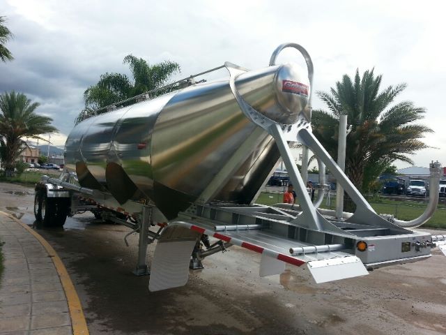 Pneumatic dry bulk trailer with a polished aluminum cylindrical tank on a V-frame hitch. Dual axles with black tires, rearmost wheels visible. Tow hitch and support legs extend under tank; rear apron and reflective tape.