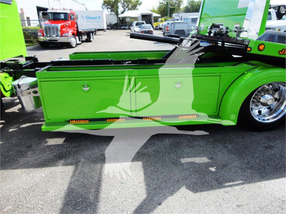 Green Peterbilt truck side with storage compartments and chrome wheel, visible hydraulic components and towing equipment.