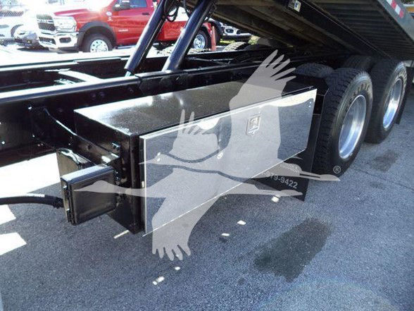 Truck trailer undercarriage showing a rectangular metal toolbox/box mounted near the wheel, with a hitch receiver and a black frame; visible dual rear wheels and a black surface.