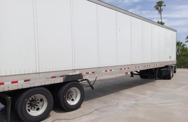 Trailer side view showing a long white cargo trailer with twin rear axles, reflective tape, and a black undercarriage.