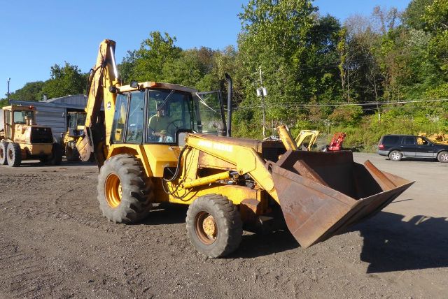John Deere 710B backhoe loader in a dirt lot with bucket attached, side view, with trees in background.