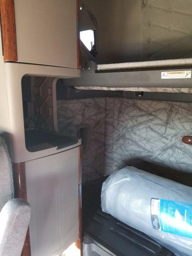 Cab interior of a Freightliner Cascadia showing a passenger side step area, with a gray fabric seat, wooden trim, and a rolled blue item (likely mattress) on the floor.