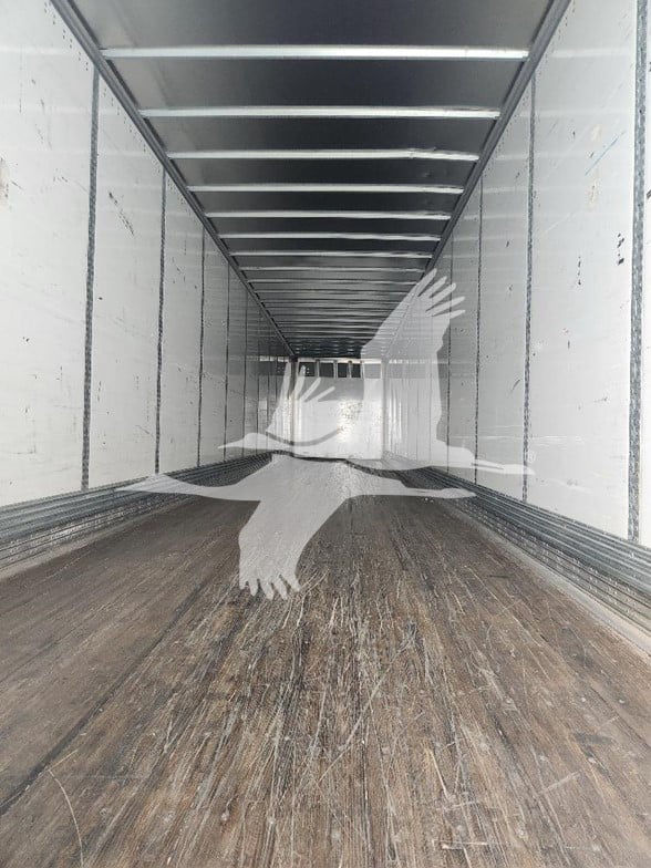 Interior of a trailer van cargo space showing wooden floor and white walls with ceiling beams.