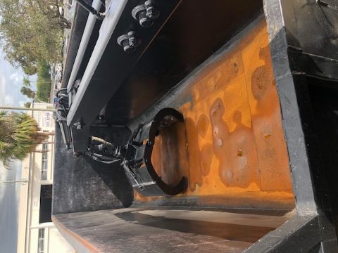 Forklift-style grapple attachment in a dump truck bed with orange rusty floor and hydraulic claw visible.