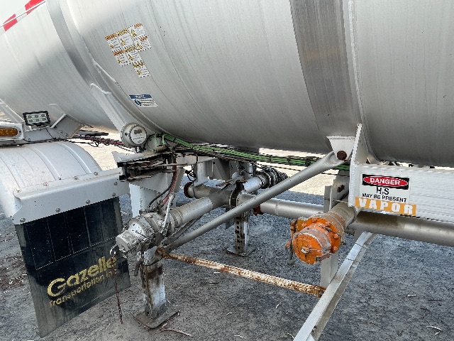 Trailer frame with cylindrical tank, hoses, and control valves; orange gearbox, warning labels, and Gazelle Technologies component; surrounding ground and support stands.
